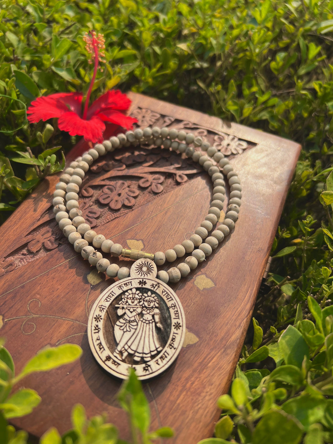 Tulsi mala with round carved locket on a wooden board beside a red hibiscus flower