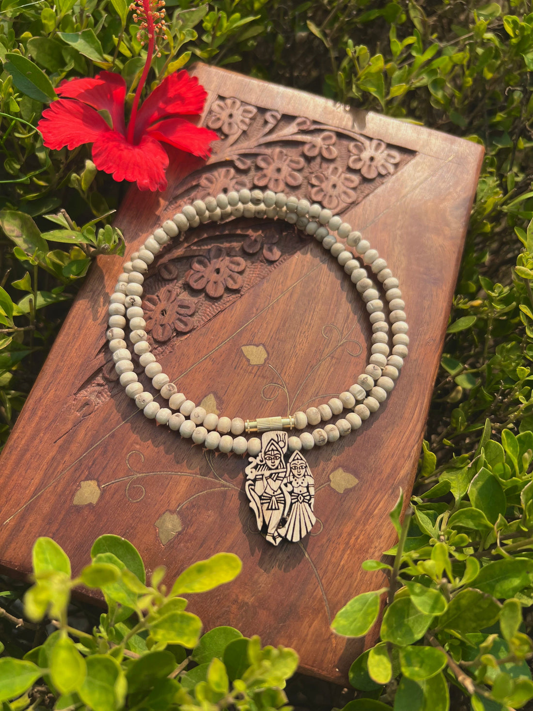 Tulsi kanthi mala with carved Radha Krishna pendant on a wooden board in greenery