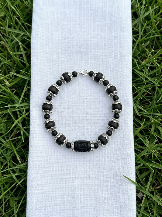 A beaded bracelet with black beads, silver spacer beads, and a central black rectangular charm, displayed on a white fabric sleeve over a green grass background.