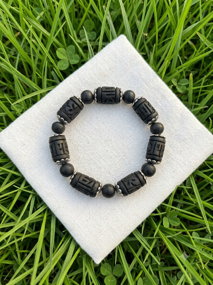 A beaded stretch bracelet with matte black cylindrical beads and small round black beads, displayed on a white fabric square over green grass.